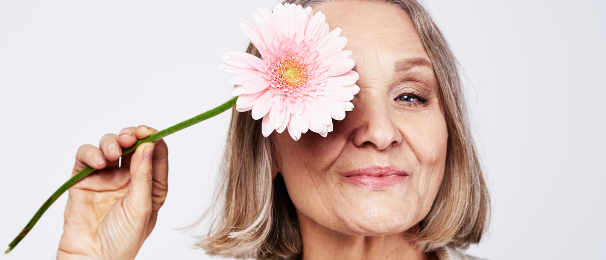 Donna sorridente in menopausa che tiene un fiore rosa davanti a un occhio, mostrando la pelle luminosa e i bei capelli dopo aver assunto collagene da bere