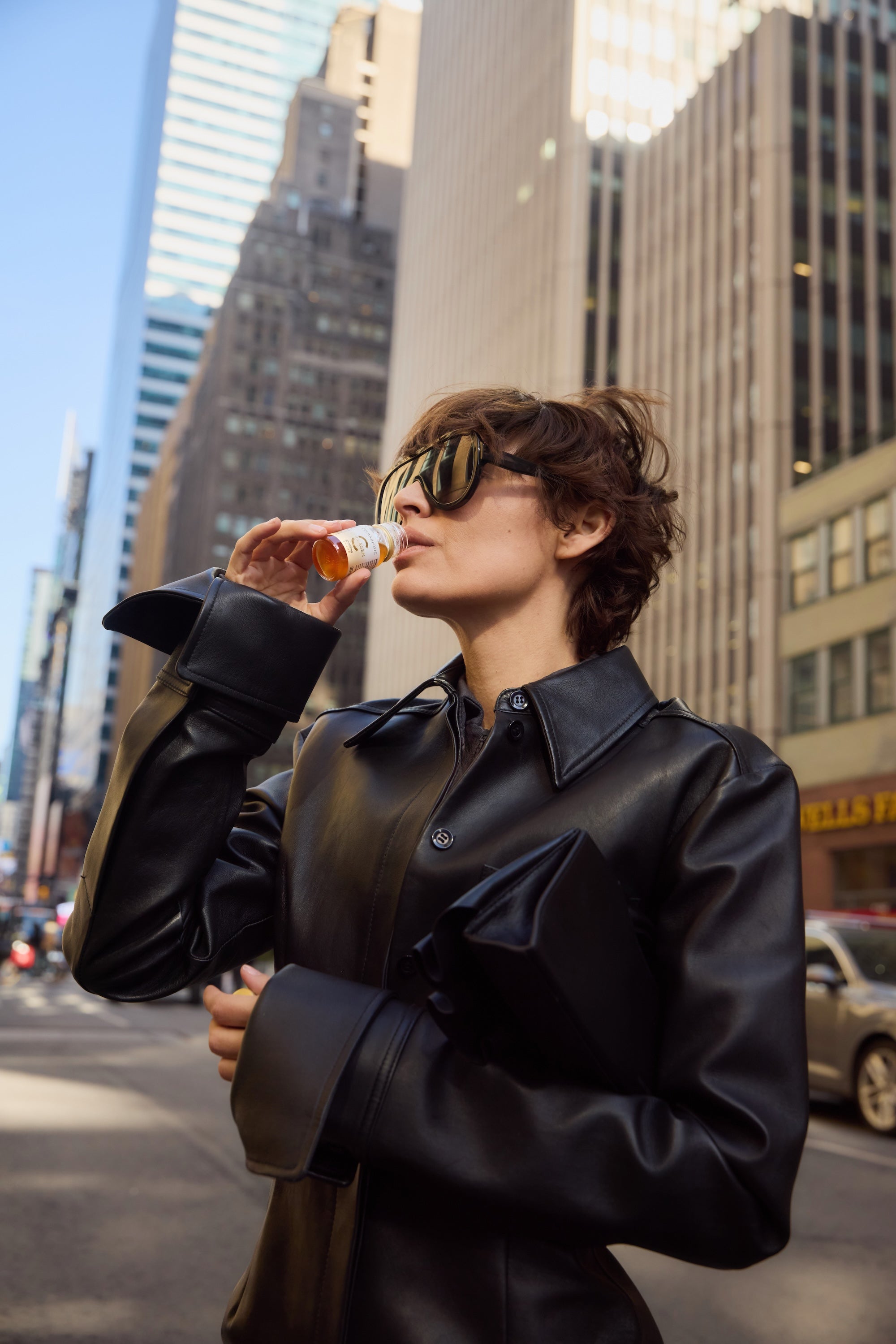 donna con occhiali da sole e cappotto nero beve un flaconcino di Primabiotic Collagen tra i grattacieli della città
